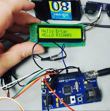 A hand holding an LCD screen displaying greetings, connected to a microcontroller board with wires.