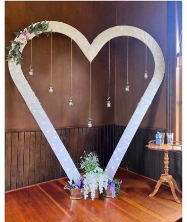 Large heart-shaped frame decorated with lights, flowers, and hanging jars in a cozy wooden room.