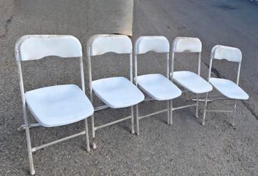 Five white folding chairs connected in a row on pavement.