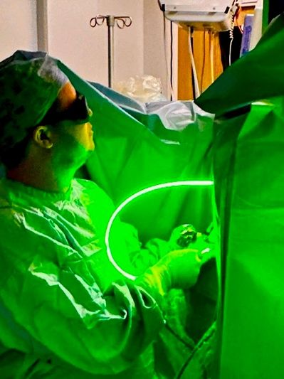 Surgeon performs laser surgery with protective eyewear in a green-lit operating room.