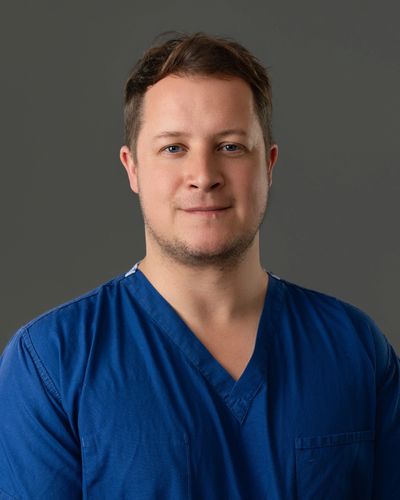 Portrait of a man in blue medical scrubs against a gray background.