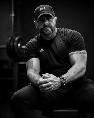 Muscular man in a gym, wearing a cap and Adidas shirt, sitting and looking serious.