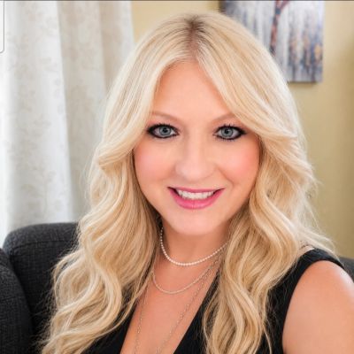 Smiling blonde woman with wavy hair and pearl necklace.