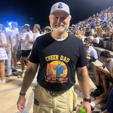 Smiling man in "Cheer Dad" T-shirt at a lively nighttime sports event.