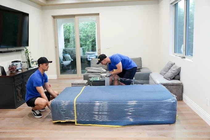 Two men wrapping a mattress with protective plastic in a living room.