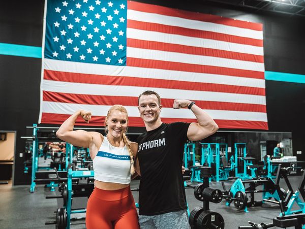 Two fit individuals flex muscles in a gym with a large American flag behind.