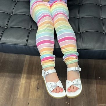 Child wearing rainbow-striped pants and white sandals sitting on a black couch.