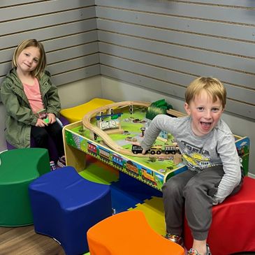 Two children playing happily with a train set on a colorful table surrounded by stools.