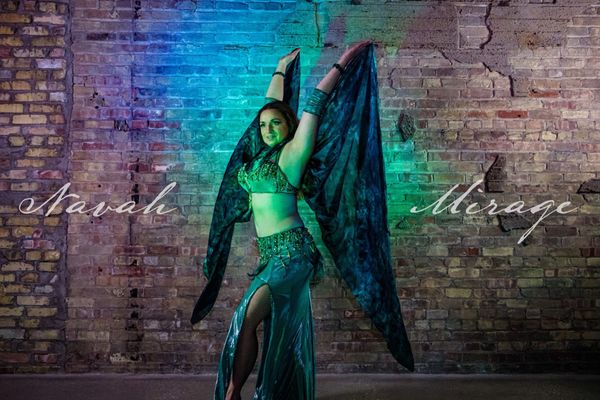 Woman in blue belly dance costume poses against a brick wall with 'Navah Mirage' text.