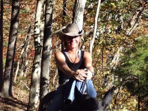 Smiling woman wearing a cowboy hat riding a horse in a sunlit forest.