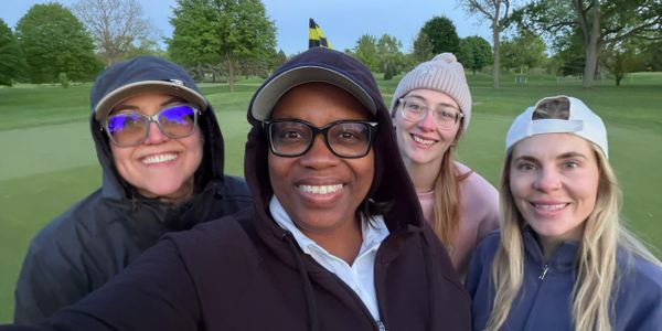 Four women smiling together outdoors on a golf course.