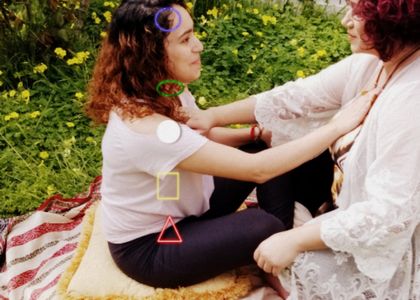 A student  shown with the 5 elements chakra points in the energy body and the practitioner placing her arms on the heart chakra. This is outside on the lawn with many yellow flowers. 
