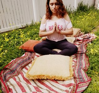 A student shown in the meditation position sitting outside on the grass with pillows during the day.