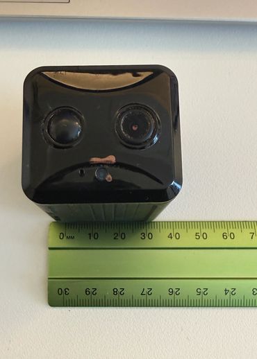 Black rectangular device with two lenses, placed next to a green ruler.
