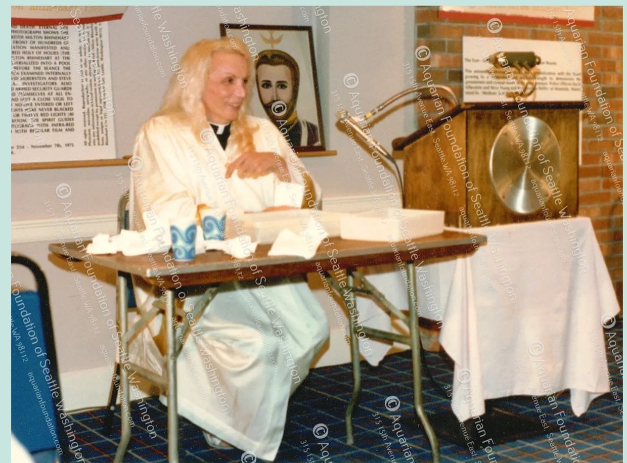 Person with long blonde hair sitting at a table in a room with framed artwork and a podium.