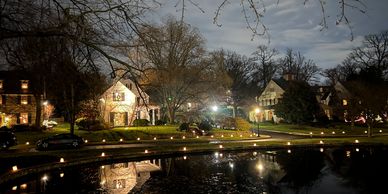 A quiet neighborhood at night with houses reflected in a pond and lights lining the street.