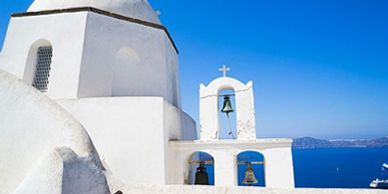White greek-looking church with cross, three bells, and ocean in the background