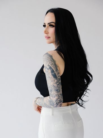 Woman with long black hair and a detailed sleeve tattoo in a black top and white pants.