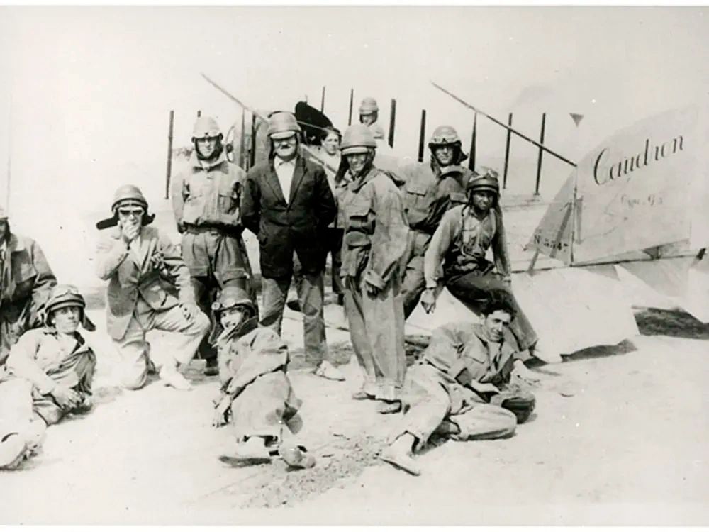 Group of early aviators posing beside a vintage biplane named 'Caindon'.