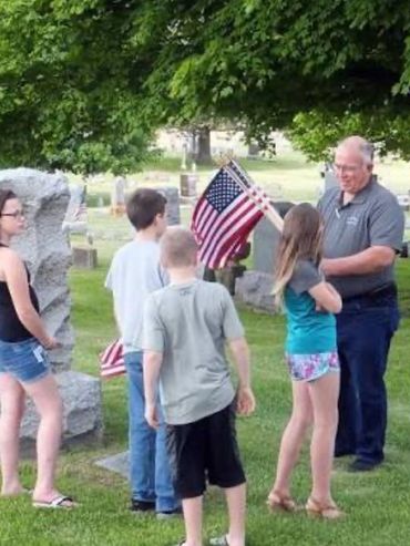 An elderly man and children at a cemetery with American flags.