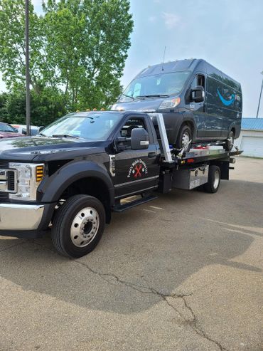 Tow truck carrying a dark Amazon Prime delivery van on its flatbed.