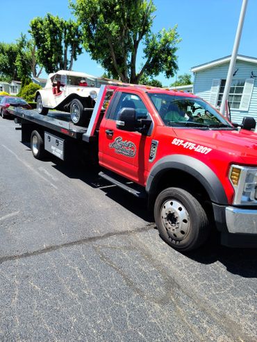 Red tow truck carrying a vintage white car on a sunny day.