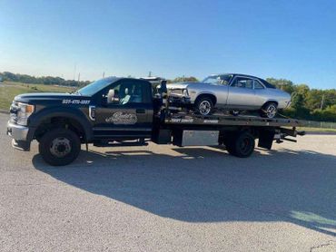 A flatbed tow truck carrying a classic silver car on a sunny day.