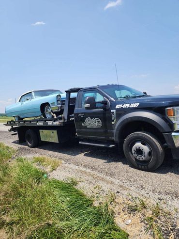 A black tow truck carrying a light blue vintage car on a sunny day.