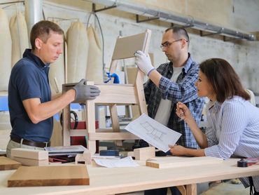 Three people assembling a wooden chair using blueprints in a workshop.