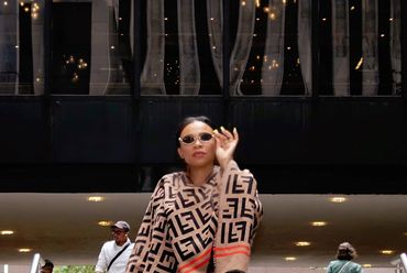 A stylish woman in sunglasses and patterned shawl walks confidently in an urban setting with stairs and people around.