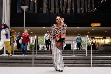 A stylish woman in sunglasses and patterned shawl walks confidently in an urban setting with stairs and people around.