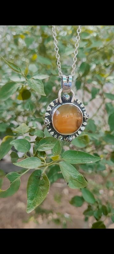 Silver pendant necklace with an orange gemstone, hanging among green leaves.