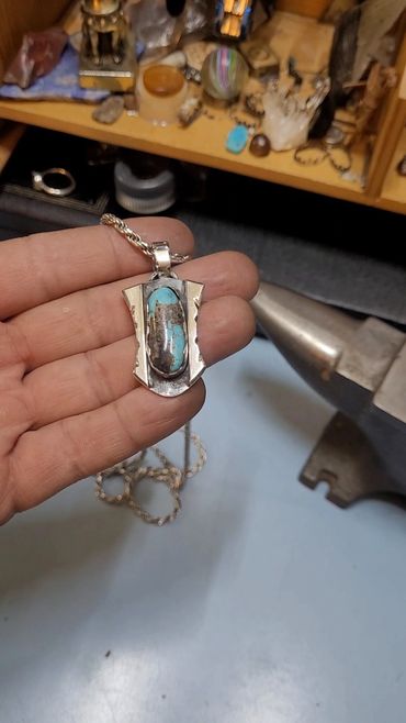 Hand holding a handcrafted silver and turquoise pendant with a chain.
