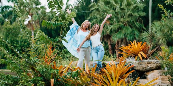 Two women joyfully posing with arms raised in a lush tropical garden.