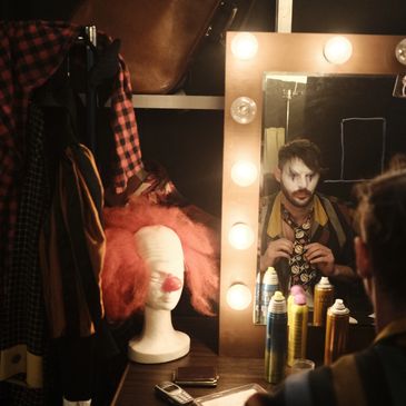 Clown schminkt sich vor Spiegel mit Perücke nebenan.