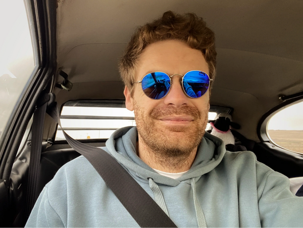 Man wearing reflective blue sunglasses and a hoodie, smiling inside a car with a seatbelt on.