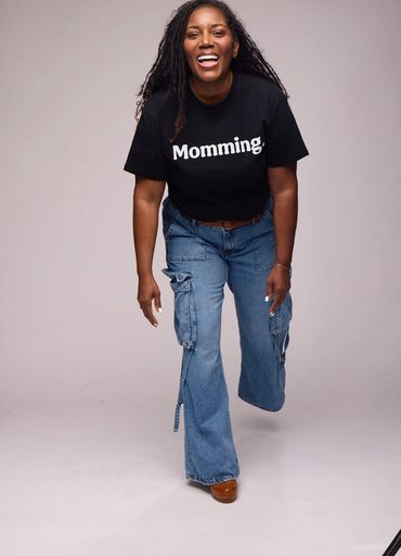Woman smiling wearing a black 'Momming.' t-shirt and blue jeans.