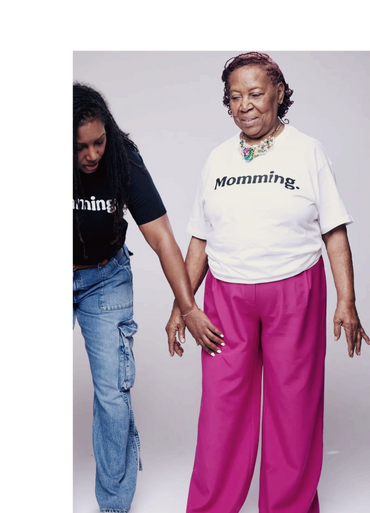 Two women holding hands, wearing 'Momming.' shirts, sharing a tender moment.
