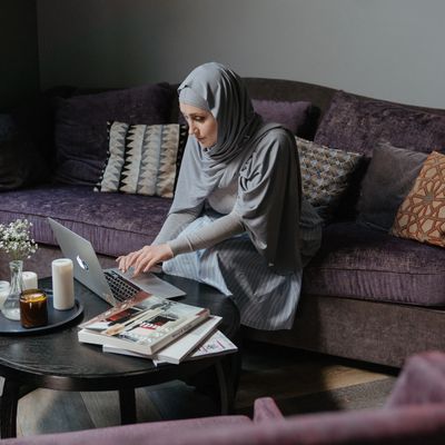 Woman wearing a hijab, sitting on a couch and working on a computer.