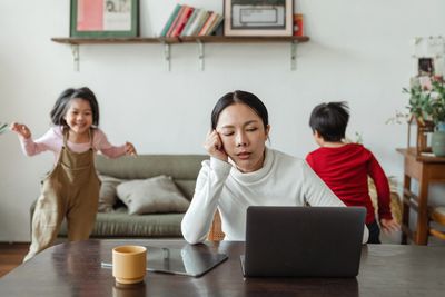 Tired, asian woman in front of a computer with children running around in the background