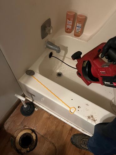 Using a drain snake to unclog a bathtub drain with cleaning supplies nearby.