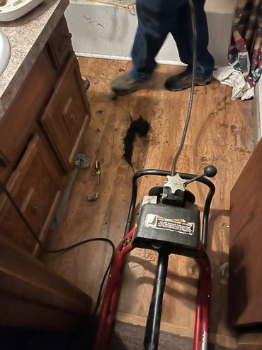 A person using a drain cleaning machine on a wooden floor with hair debris scattered.