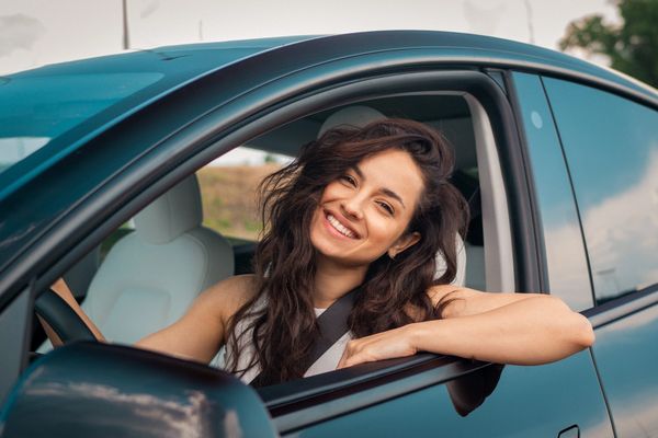 A woman with long, wavy hair leans out of a car window, smiling, while wearing a seatbelt