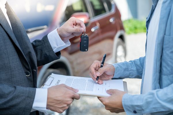 A person in a suit hands over car keys while the buyer signs a contract with a pen
