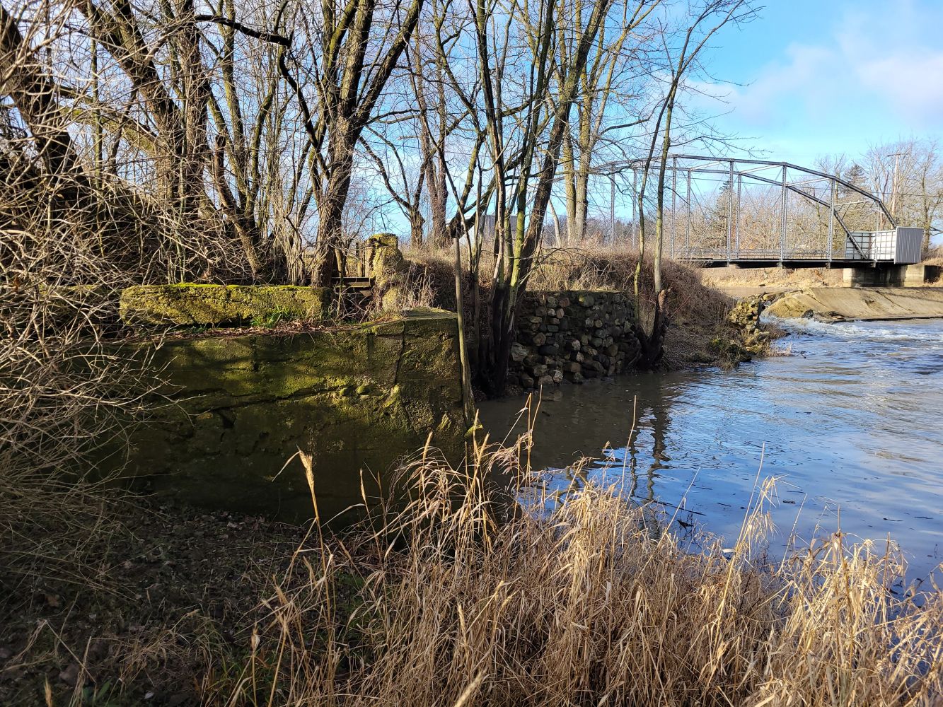 Riverbank with bare trees, a metal bridge, and mossy stone walls on a sunny day.
