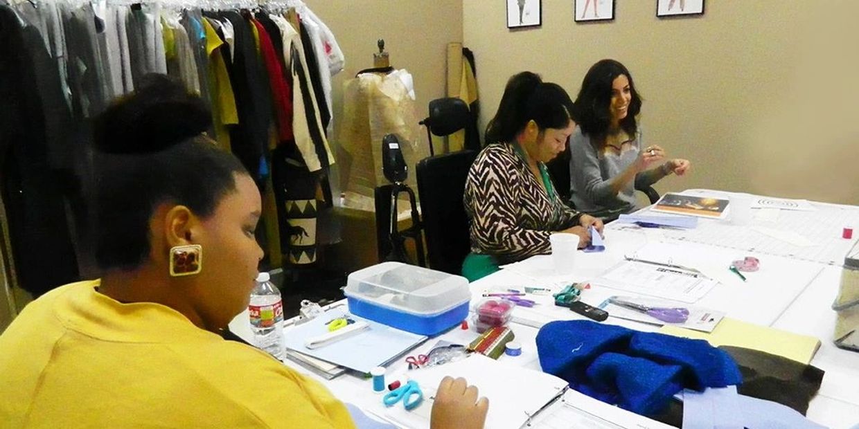 Three women working on crafts at a table in a room with clothes.