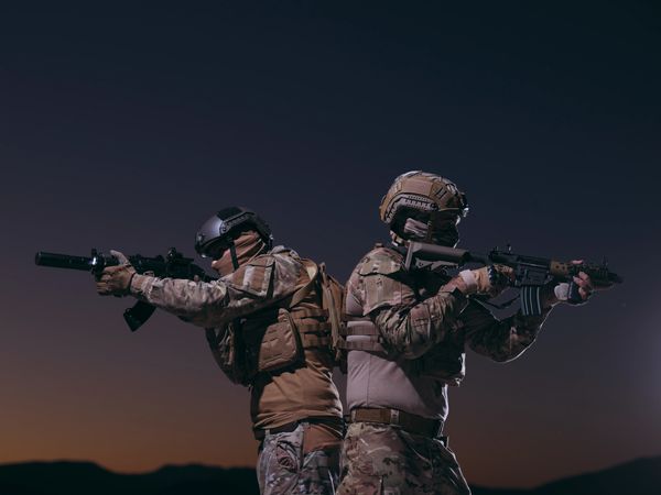 Two figures in camouflage gear and helmets holding rifles back-to-back at dusk.