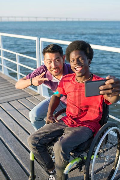 Two friends, one in a wheelchair, taking a joyful selfie by the water on a sunny day.