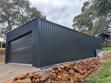 Newly constructed black metal garage with a roller door, surrounded by trees and natural landscaping.