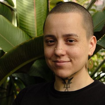 Person with shaved head, facial piercings, and tattoos smiling outdoors.